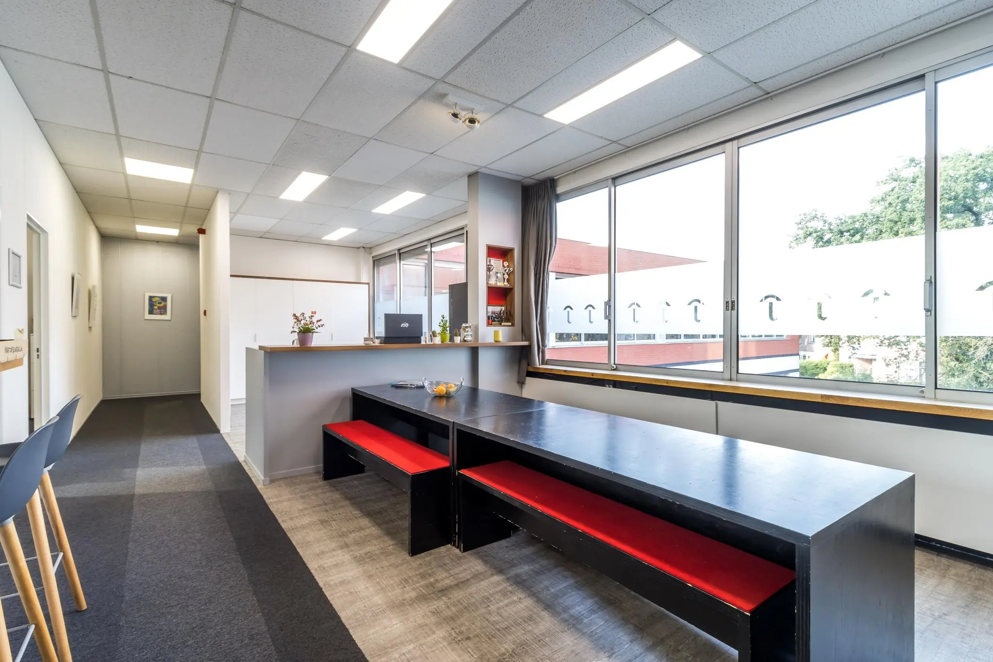 Moderne kantineruimte aan de Jan van Eycklaan met lange zwarte tafel, rode banken en grote ramen met uitzicht op buiten.